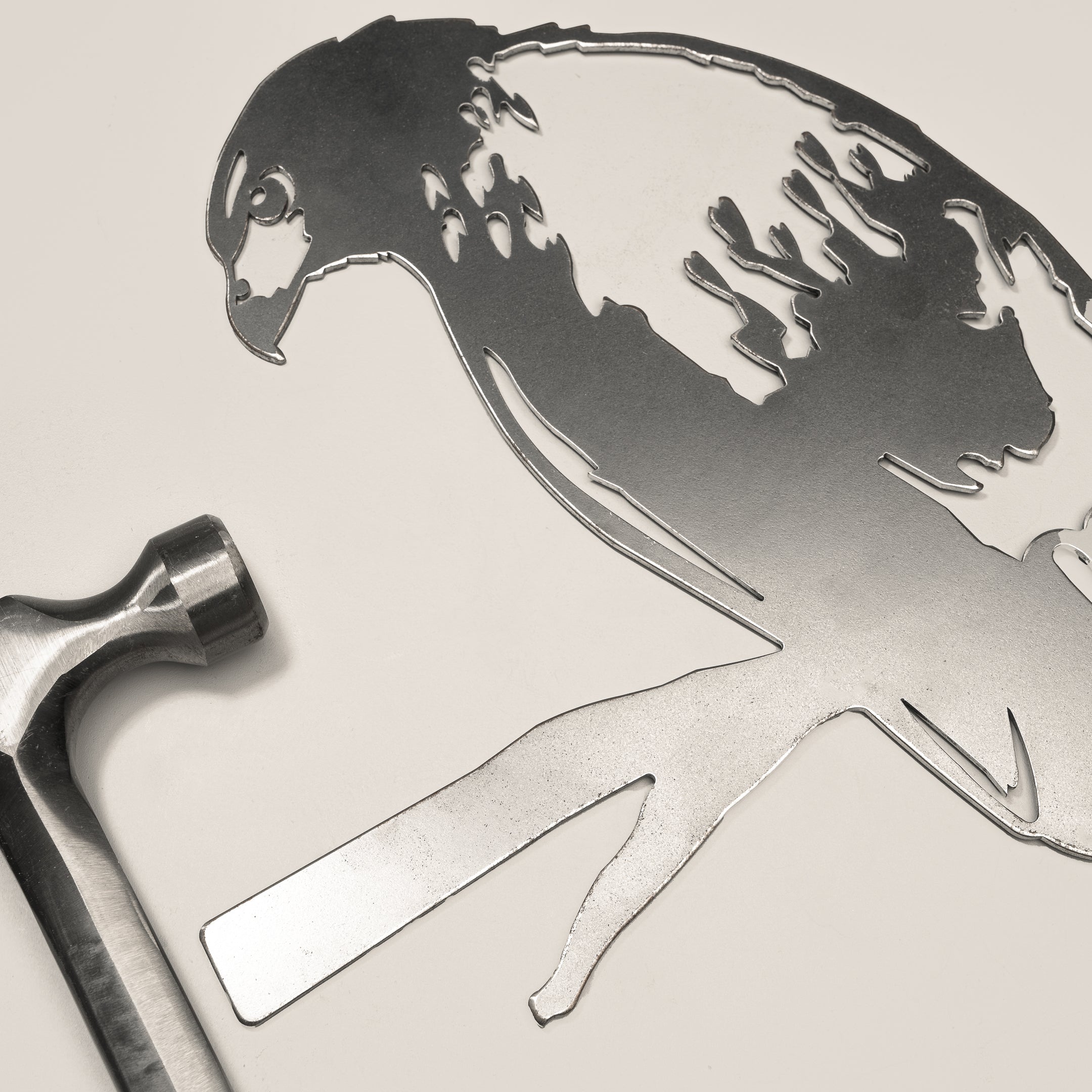 A Red-Tailed Hawk metal silhouette by Metalbird Canada lies next to a metal hammer on a light surface. This detailed yard art makes a unique gift and adds character to any outdoor space. The hammer is positioned left of the artwork.