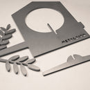 Gray metal pieces from the Metalbird Canada Bird Feeder, including a house-shaped silhouette with METALBIRD text, two leafy branches, and a curved piece, are arranged on a light surface.