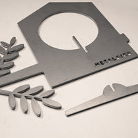 Gray metal pieces from the Metalbird Canada Bird Feeder, including a house-shaped silhouette with METALBIRD text, two leafy branches, and a curved piece, are arranged on a light surface.