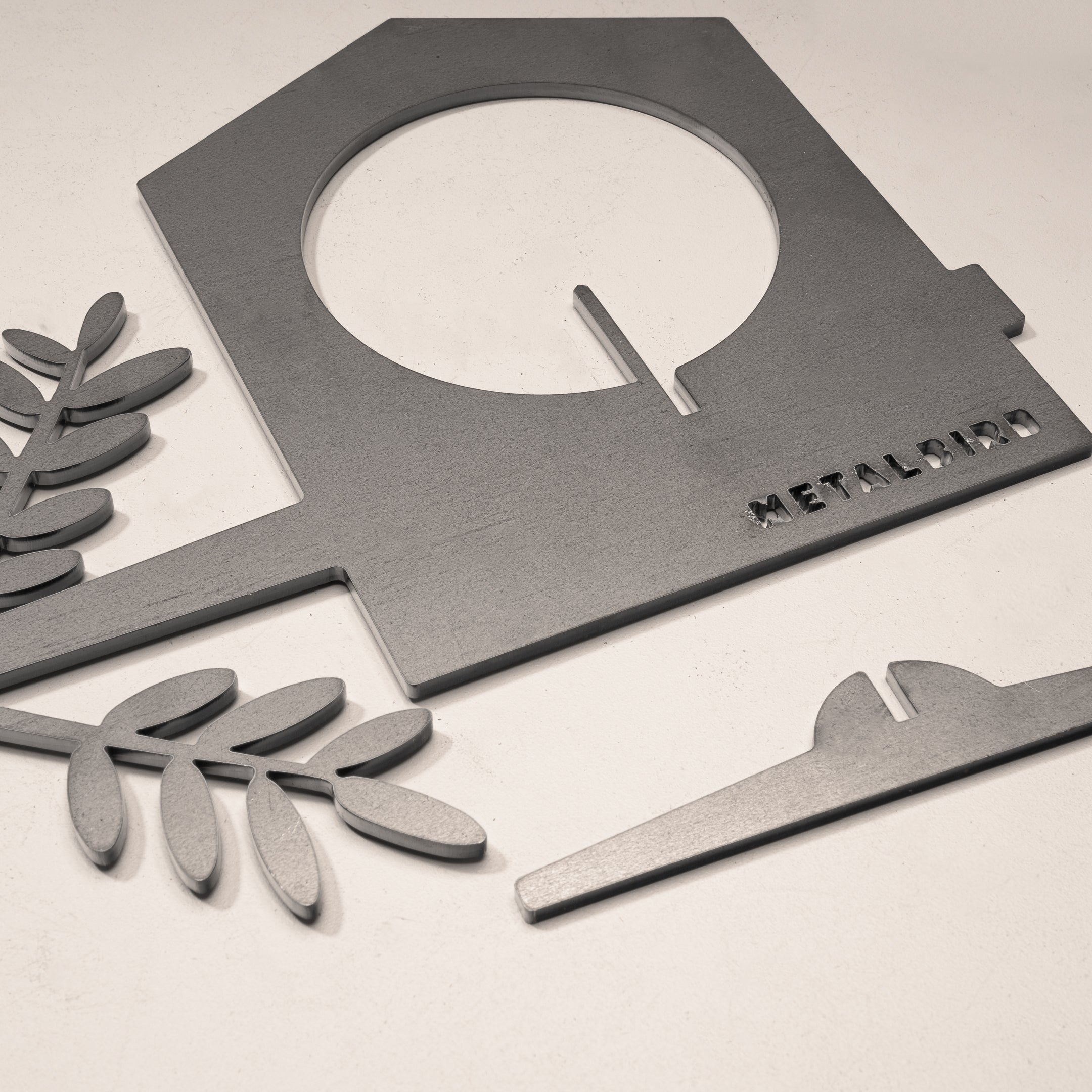 Gray metal pieces from the Metalbird Canada Bird Feeder, including a house-shaped silhouette with METALBIRD text, two leafy branches, and a curved piece, are arranged on a light surface.