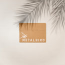 A brown envelope with a white illustration of a hummingbird on a branch and "Metalbird Canada" rests on a light surface, hinting at the unique corten steel Hummingbird inside, as palm leaf shadows cross the top right corner.