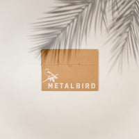 A brown envelope with a white illustration of a hummingbird on a branch and "Metalbird Canada" rests on a light surface, hinting at the unique corten steel Hummingbird inside, as palm leaf shadows cross the top right corner.
