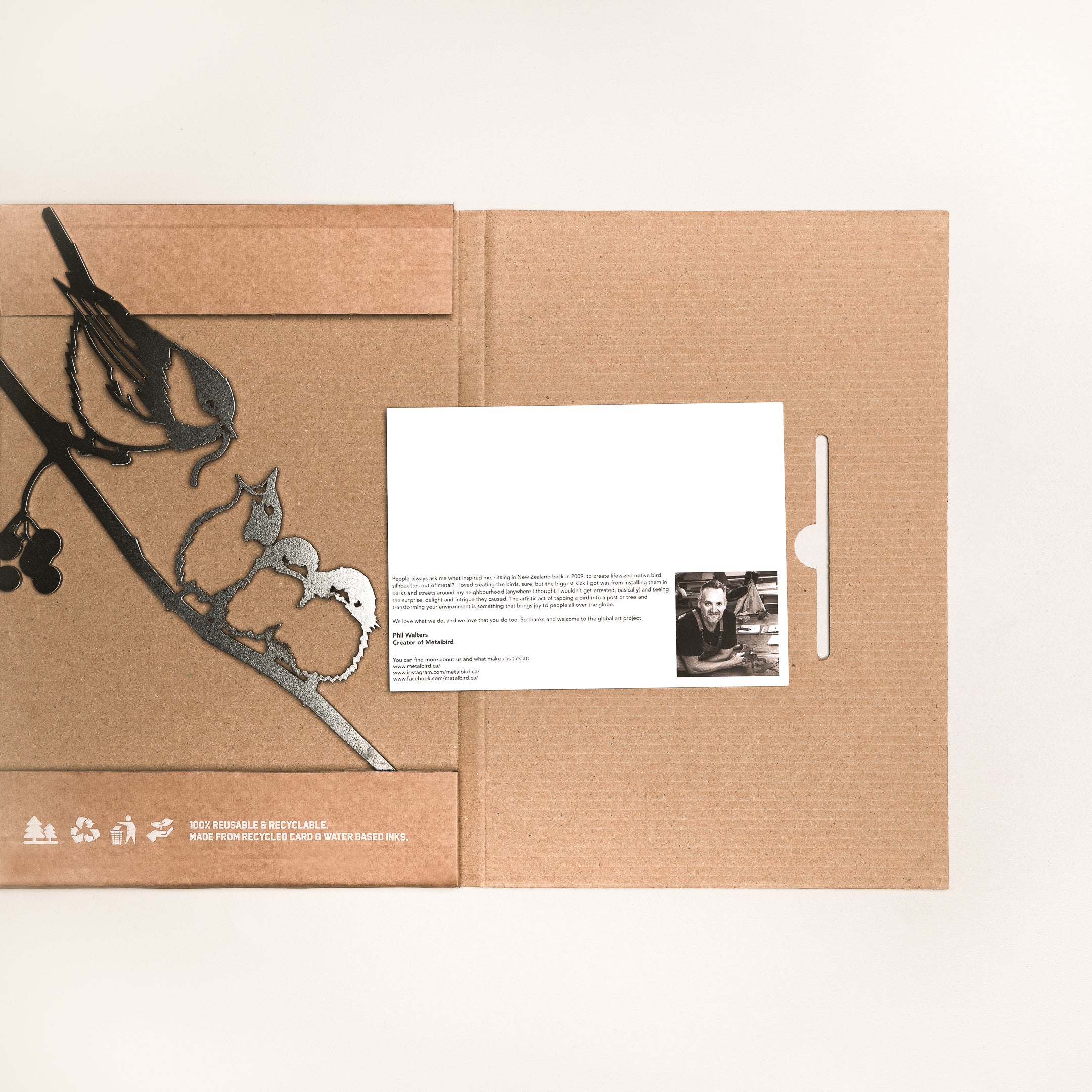 The Chickadee & Chicks by Metalbird Canada is a brown cardboard folder featuring a black floral and bird silhouette, partially open to reveal a postcard with a black-and-white photo and text—perfect as a thoughtful gift.