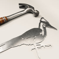 The Metalbird Canada Pileated Woodpecker metal silhouette rests on a light surface beside a hammer with a wooden handle and metal head—ideal for adding unique yard art to your outdoor space.