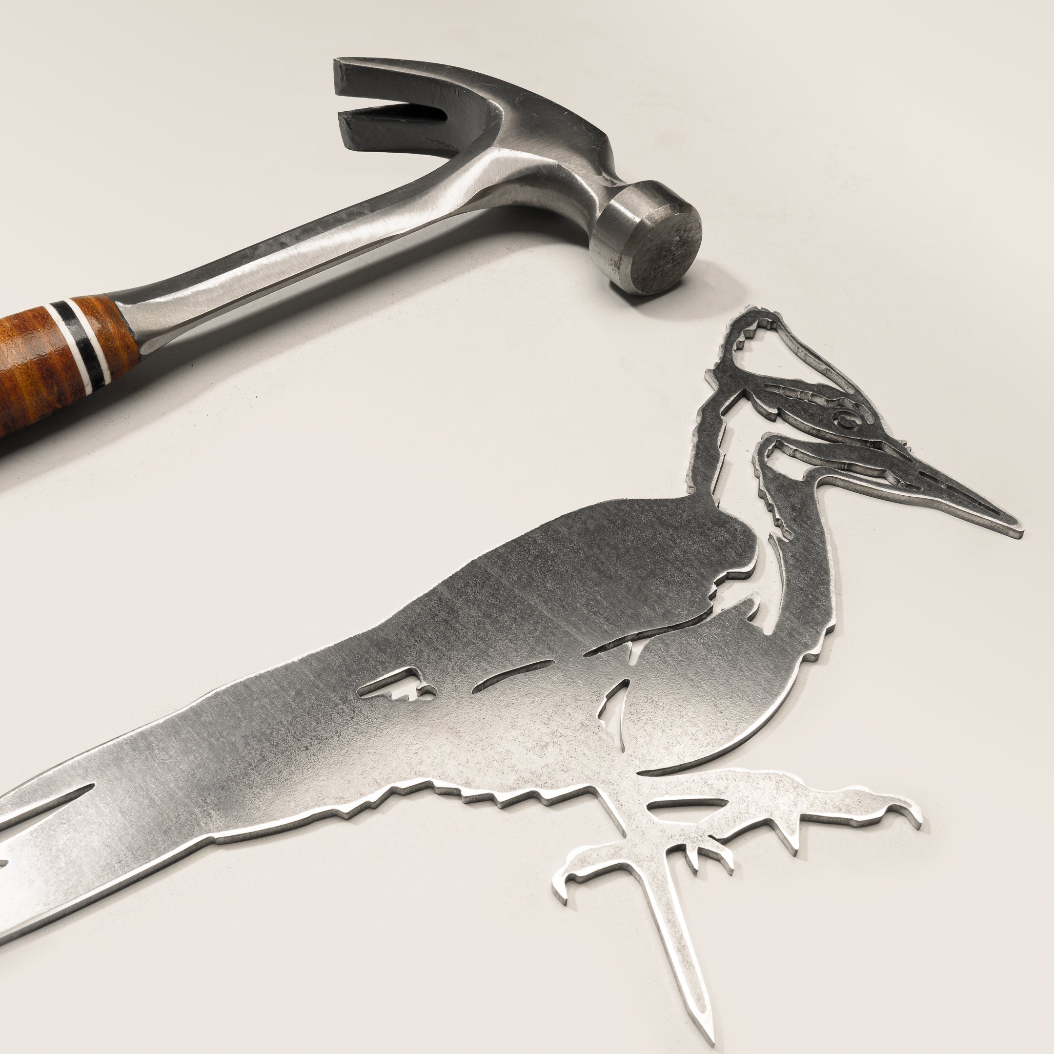 The Metalbird Canada Pileated Woodpecker metal silhouette rests on a light surface beside a hammer with a wooden handle and metal head—ideal for adding unique yard art to your outdoor space.