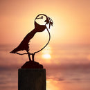 The Puffin by Metalbird Canada perches on a post as eye-catching yard art, its corten steel form set against a glowing ocean and warm sunset.