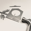 A flat metal bird feeder from Metalbird Canada, shaped like a birdhouse with a branch and leaves, lies on a white surface beside a hammer and metal crossbar—ideal as yard art or a thoughtful gift.