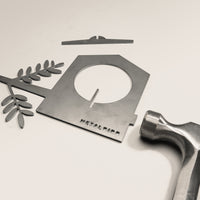 A flat metal bird feeder from Metalbird Canada, shaped like a birdhouse with a branch and leaves, lies on a white surface beside a hammer and metal crossbar—ideal as yard art or a thoughtful gift.