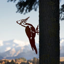 The Metalbird Canada Pileated Woodpecker metal silhouette is mounted on a tree trunk, making bold yard art that stands out against a blurred backdrop of snow-capped mountains and blue sky.