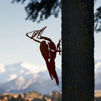 The Metalbird Canada Pileated Woodpecker metal silhouette is mounted on a tree trunk, making bold yard art that stands out against a blurred backdrop of snow-capped mountains and blue sky.