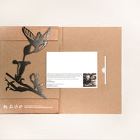 The "Hummingbird" folder by Metalbird Canada features a corten steel hummingbird and flower design on brown cardboard, partially open to show a white card with printed text and a small black-and-white portrait inside.