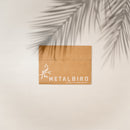 A brown cardboard box labeled Metalbird Canada with a bird graphic rests on a light surface shaded by a palm leaf—hinting at the Stick Bird Feeder, unique garden art or silhouette yard art, inside.