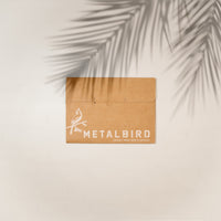 A brown cardboard box labeled Metalbird Canada with a bird graphic rests on a light surface shaded by a palm leaf—hinting at the Stick Bird Feeder, unique garden art or silhouette yard art, inside.