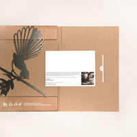 The Fantail / New Zealand Pīwakawaka by Metalbird Canada features a brown cardboard folder with a black corten steel bird silhouette on the left and a white card with text and photo on the right—an ideal unique gift.
