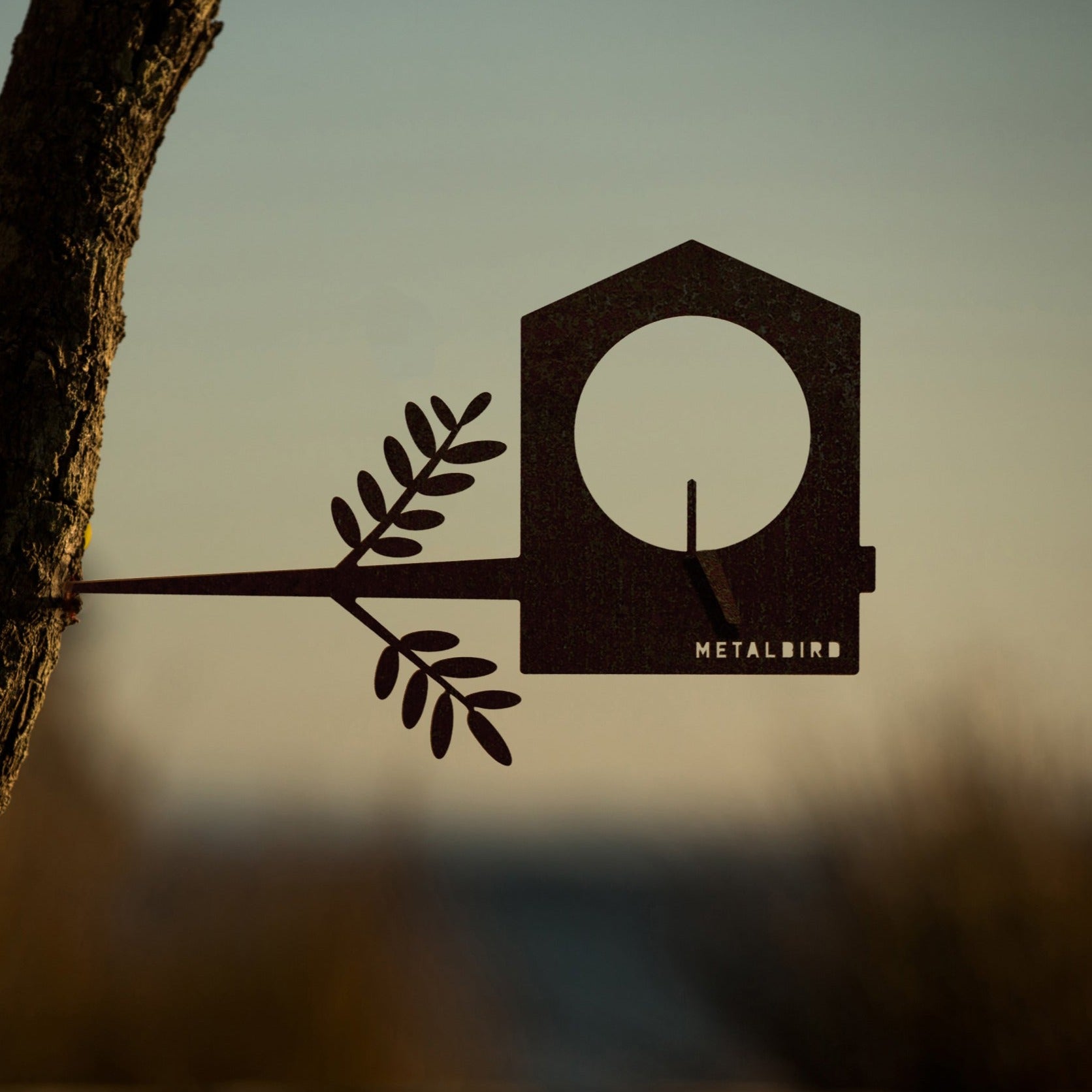 A Metalbird Canada Bird Feeder with a branch and leaf design is mounted on a tree trunk, set against a blurred sunset background. The word METALBIRD appears on this distinctive garden art piece.