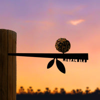 The Stick Bird Feeder by Metalbird Canada features a corten steel bird silhouette on a wooden post, holding a round seed ball. Set against a vibrant sunset and dark treetops, this charming yard art adds unique style to any garden.