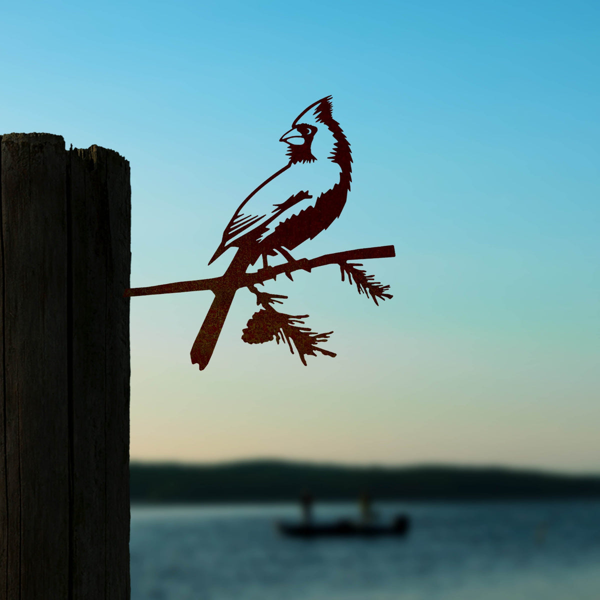 Cardinal steel silhouette on a wooden post beside a lake at sunset.