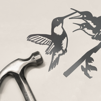 A silver hammer rests beside the limited edition Metalbird Canada "Hummingbird & Chicks" cutout, featuring two hummingbirds on a branch—an ideal Mother's Day gift set against a light background.