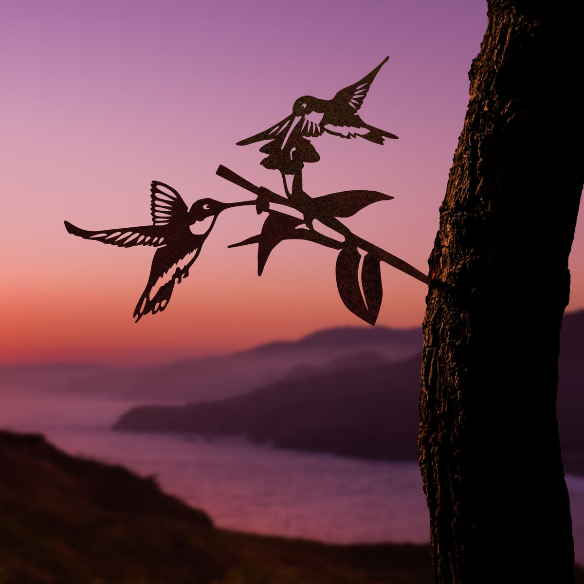 Connected hummingbirds and flower steel silhouette mounted on a tree above water at sunset.