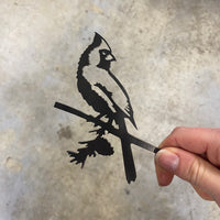 A hand holds the Mini Cardinal by Metalbird Canada, a cutout silhouette inspired by garden art in metal or corten steel, shown against a blurred gray background.