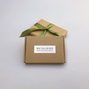 A plain brown box labeled Metalbird Canada’s Baby Bird Bundle sits before a gift wrapped in brown paper with a green ribbon—perfect for gifting unique corten steel garden art—against a light grey background.