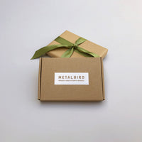 A plain brown box labeled Metalbird Canada’s Baby Bird Bundle sits before a gift wrapped in brown paper with a green ribbon—perfect for gifting unique corten steel garden art—against a light grey background.