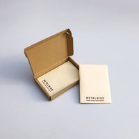 A small brown cardboard box sits open next to a closed cream-colored envelope with Metalbird Canada printed on it, displaying the Baby Bird Bundle - Hummingbird Trio, both resting on a light gray surface.