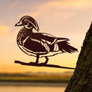 The DU Wood Duck - Large by Metalbird Canada, a striking metal bird silhouette, is mounted on a tree trunk and stands out beautifully against a glowing sunset and shimmering water backdrop.