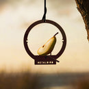 A Hanging Bird Feeder by Metalbird Canada, shaped like a bird with "METALBIRD" cut out and a pear slice for its body, hangs from a tree branch. The softly blurred background glows with warm sunset tones.
