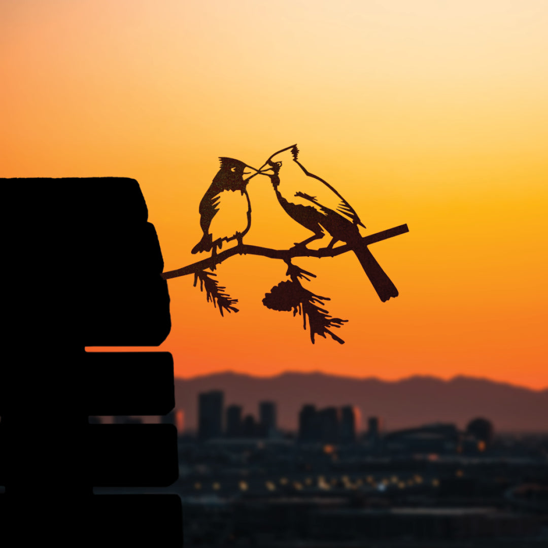 Two Cardinals meeting beak to beak in a steel silhouette against a city sunset.
