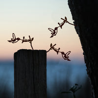 Metalbird Canada’s Baby Bird Bundle – Hummingbird Trio metal silhouettes adorn a wooden post and tree at sunset, their shapes standing out against a softly blurred background.