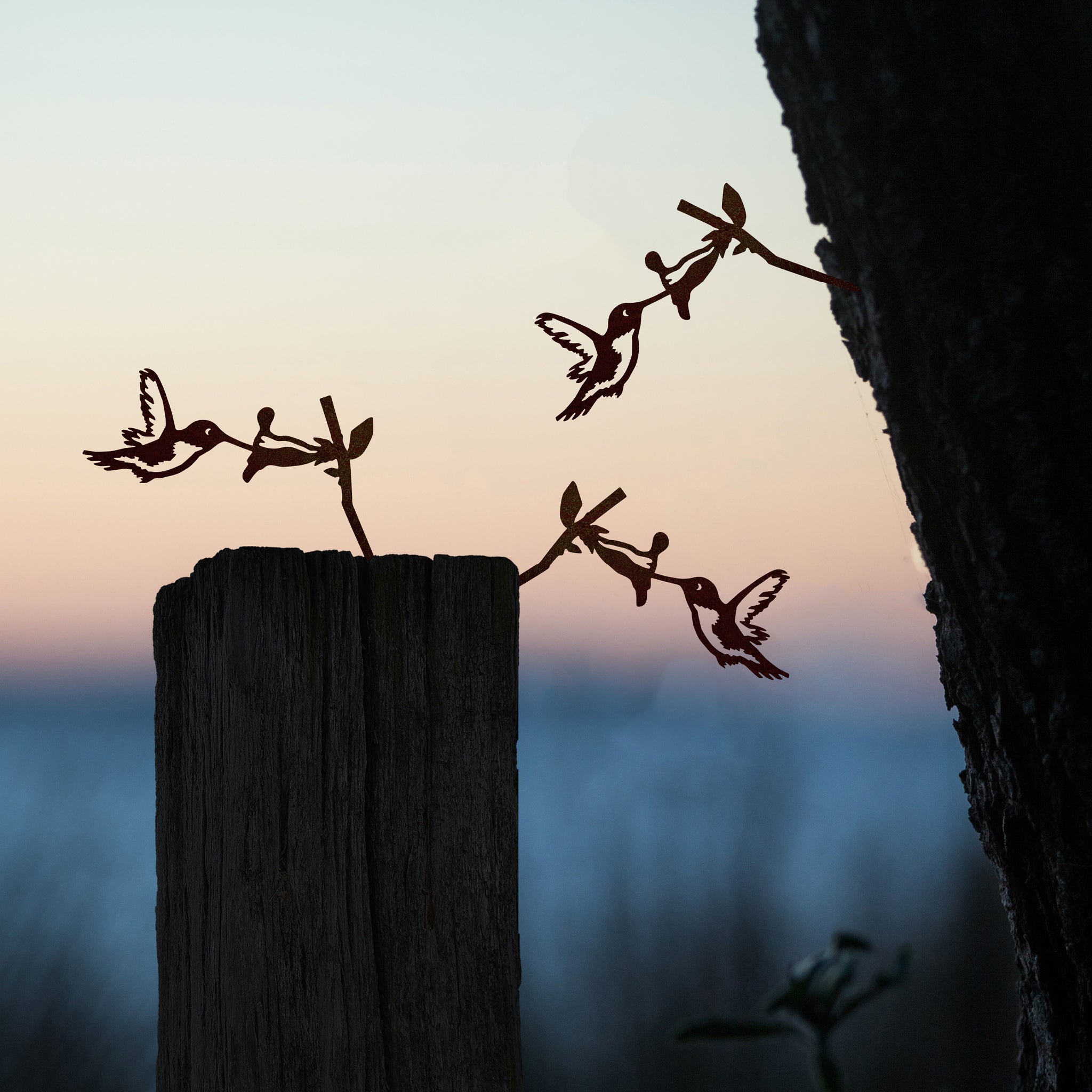 Metalbird Canada’s Baby Bird Bundle – Hummingbird Trio metal silhouettes adorn a wooden post and tree at sunset, their shapes standing out against a softly blurred background.