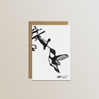 The Metalbird Canada Metalbird Greeting Card Set features a white card with a black silhouette hummingbird and flower design, inspired by metal yard art, paired with a brown envelope against a light backdrop.