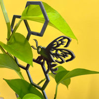 The Honeybee Plant Trellis by Metalbird Canada is displayed among green leaves, set against a vibrant yellow background.