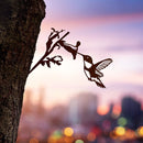 The Hummingbird by Metalbird Canada is a metal silhouette of a hummingbird feeding from a flower, mounted on a tree trunk—an ideal gift. A blurred cityscape and vibrant sunset form a stunning backdrop.