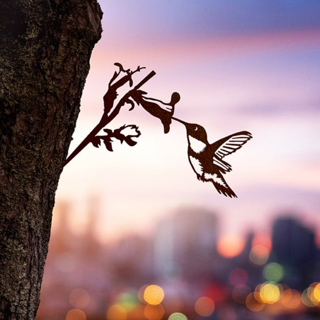 The Hummingbird by Metalbird Canada is a metal silhouette of a hummingbird feeding from a flower, mounted on a tree trunk—an ideal gift. A blurred cityscape and vibrant sunset form a stunning backdrop.