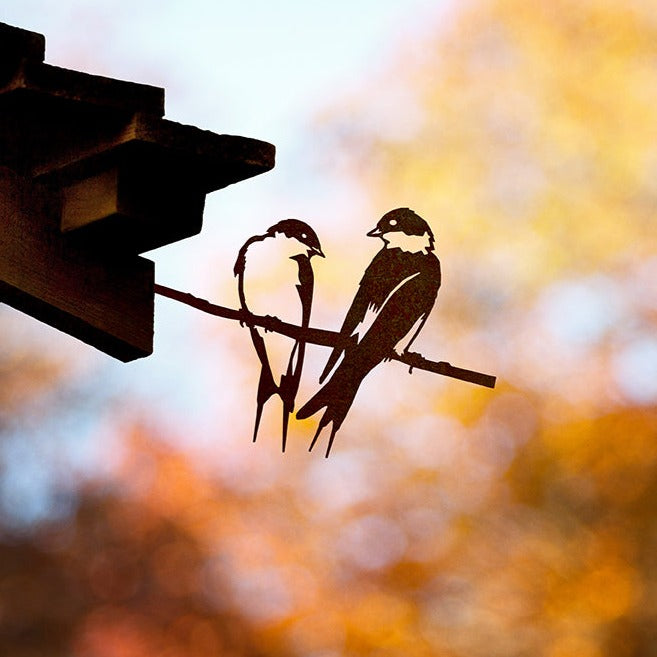 Two swallows perched together in a steel silhouette beneath wooden eaves.