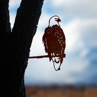 The Bald Eagle by Metalbird Canada is a corten steel silhouette of an eagle on a branch, mounted to a tree trunk to create eye-catching garden art with the sky and landscape softly blurred in the background.