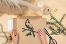 A person folds a Cardinal box from Metalbird Canada, surrounded by scissors, ribbon, dried flowers, a candle, and burlap—perfect DIY yard art inspiration on a light wooden surface.