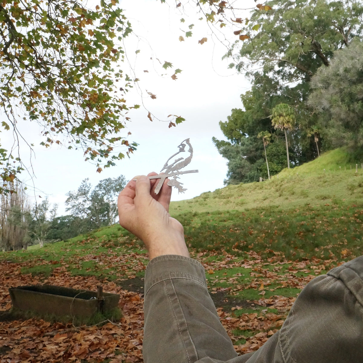 Person holding the Mini Cardinal steel silhouette outdoors among trees and fallen leaves.