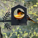A black and orange bird eats an orange slice on the Bird Feeder by Metalbird Canada, a corten steel house-shaped feeder set amid green foliage—an ideal gift for bird lovers.